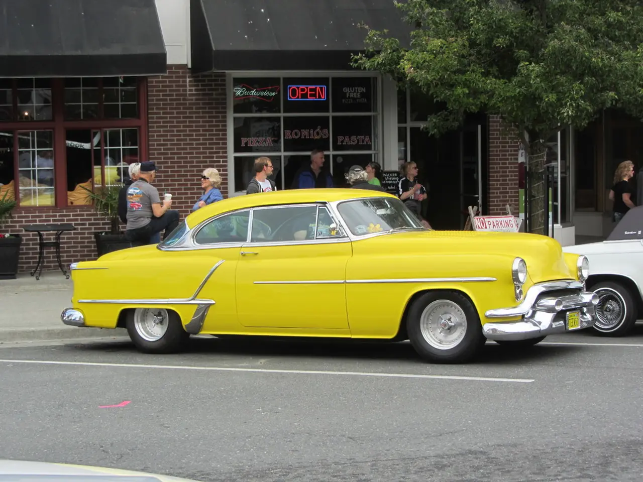 As we can see in the image there are cars, few people here and there, chairs, tree and a building.
