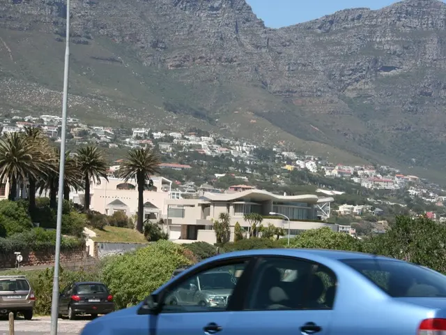 In this image there are few vehicles moving on the road, there is a pole, trees, buildings,...