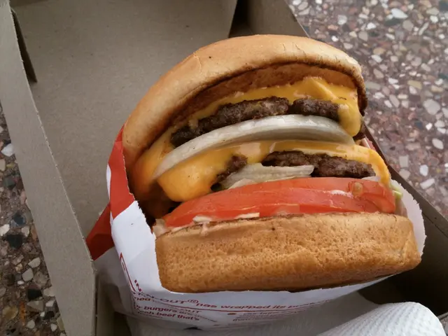 In this image I can see a burger which is brown, black, cream, orange and red in color in a paper...
