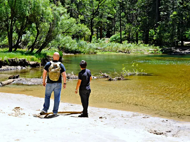 It is a river bank, on the bank there are two men standing,the first man is wearing a bag, in the...